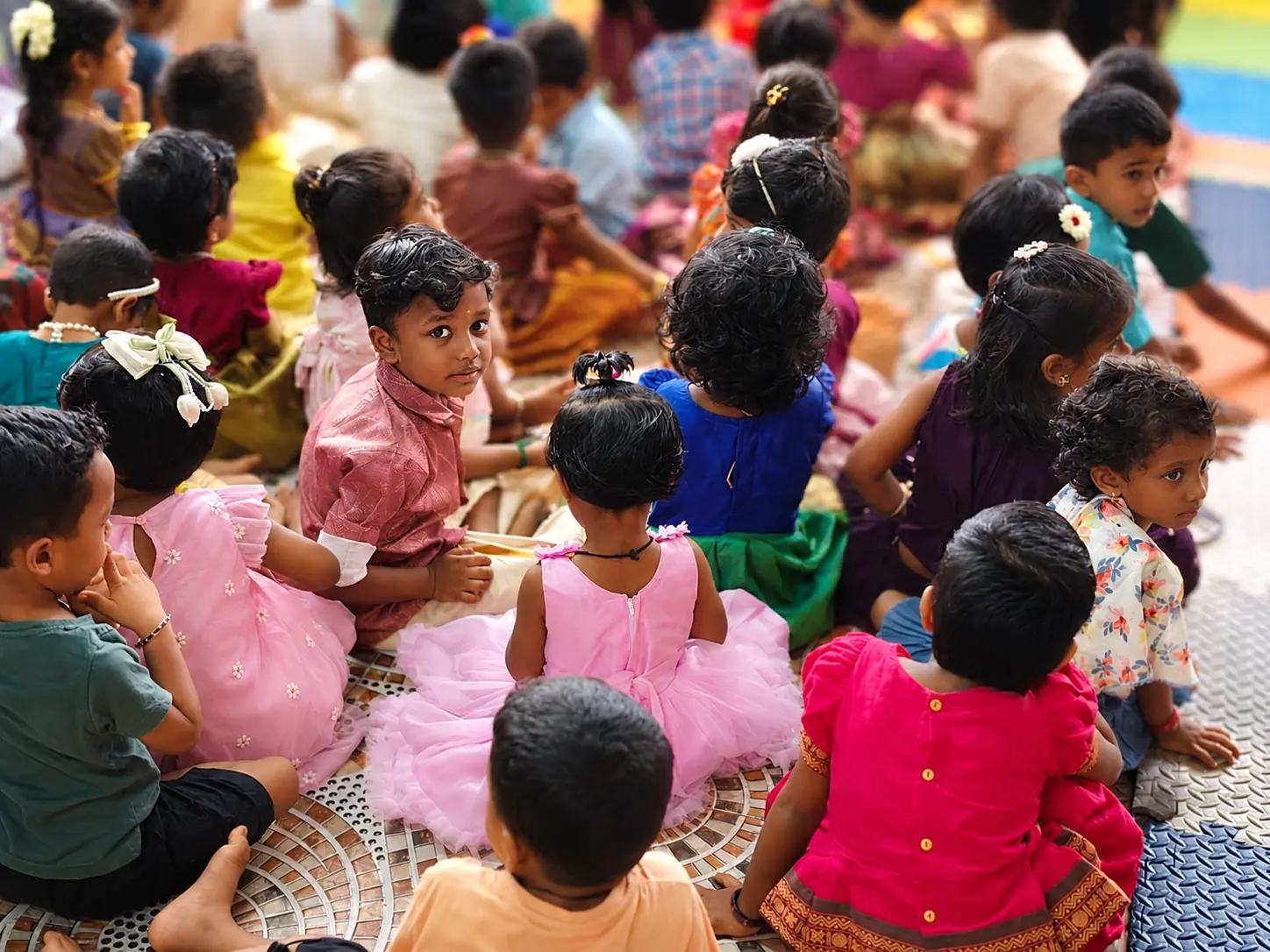 A large group of children in colourful festive outfits sitting together on the floor, seen from behind, with a few glancing at the camera