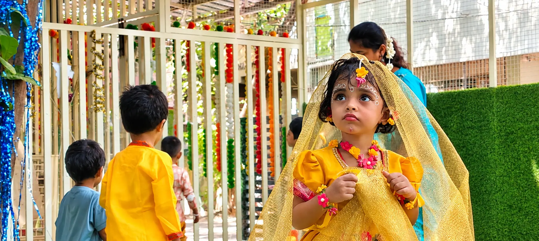 A young girl in a yellow festive dress with flower jewellery and a golden dupatta looking upward, with children and orange, red and green garlands behind her