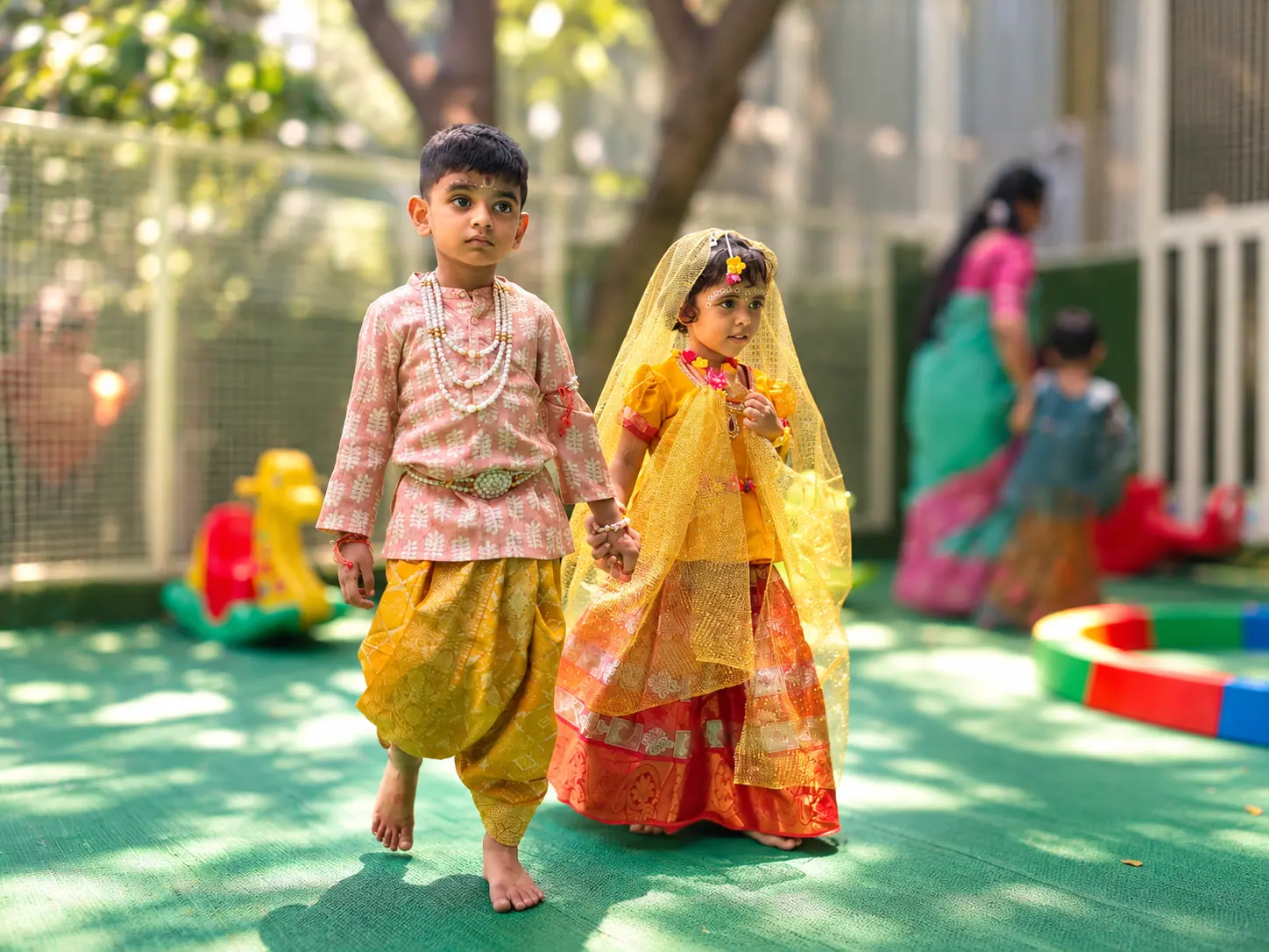 A boy in a pink printed kurta and yellow dhoti walking hand in hand with a girl in a yellow dupatta and red lehenga across the green outdoor play deck