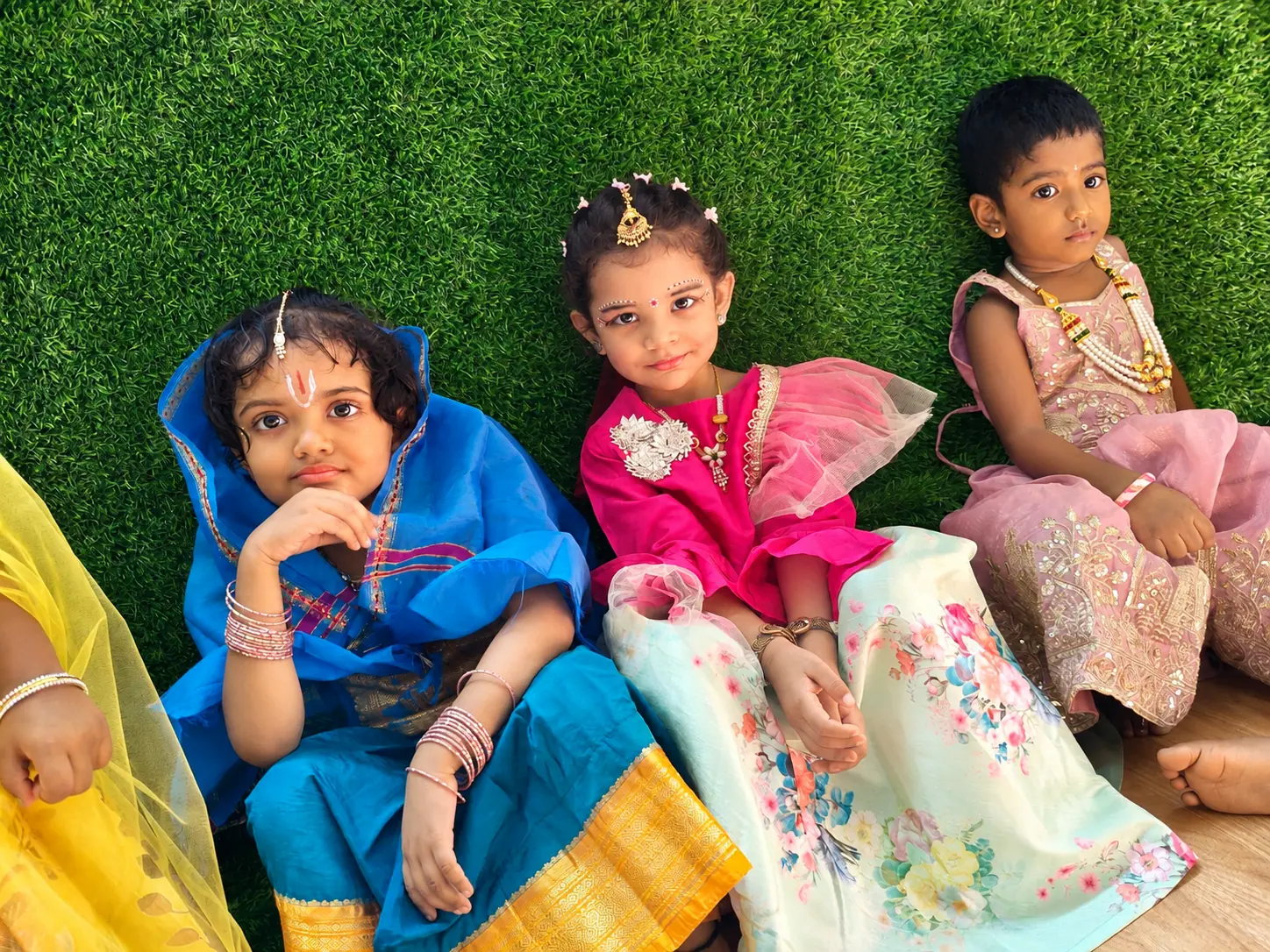 Three young girls in a blue drape and gold-bordered skirt, a pink and mint floral lehenga, and a pink embroidered outfit, seated against a green turf wall