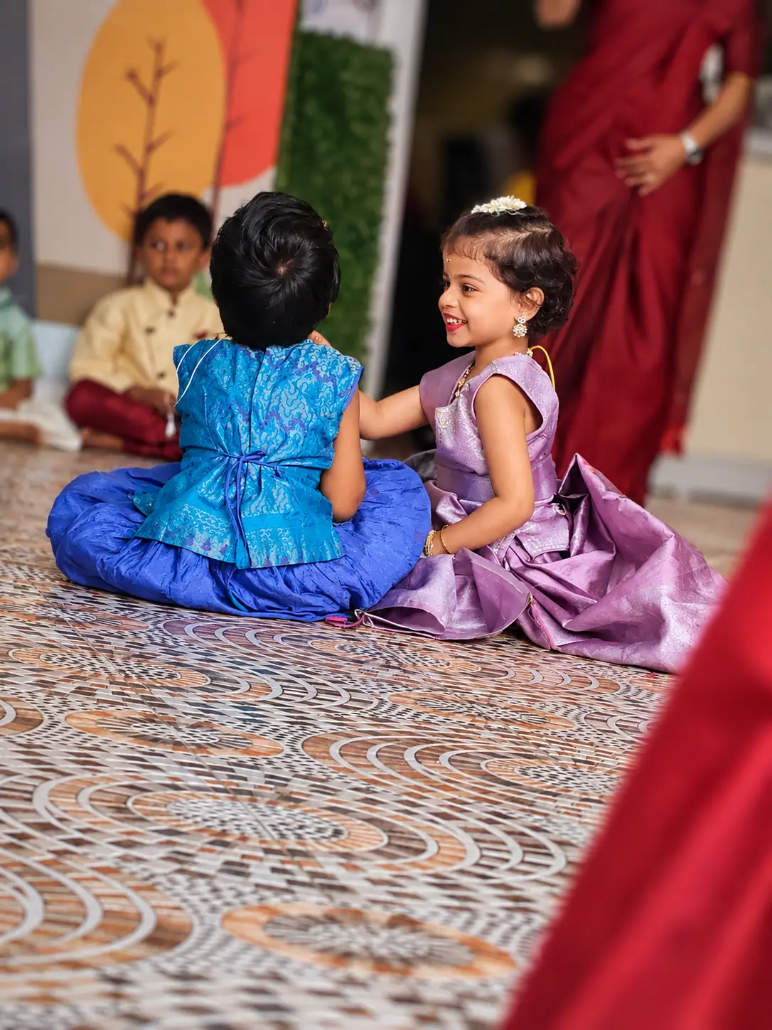 Two little girls in blue and lilac silk festive dresses sitting on a patterned floor, the girl in lilac smiling and touching the other girl's shoulder