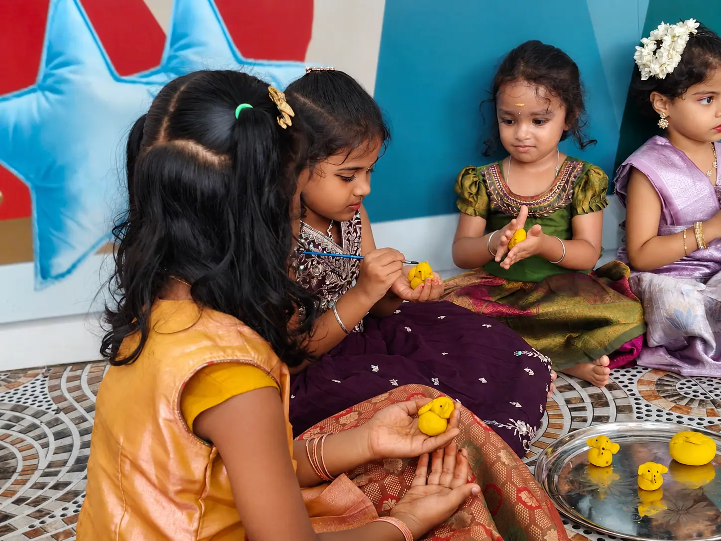 Children shaping and painting small yellow clay Ganesha figures, with finished figures resting on a steel plate