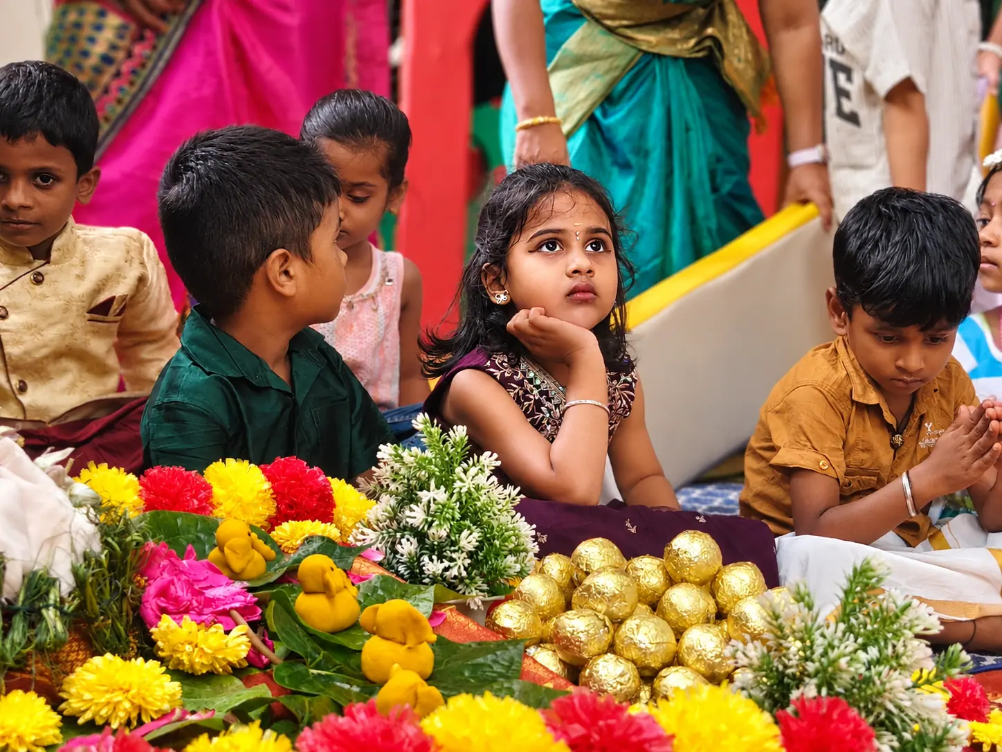 A girl in a maroon embroidered dress resting her chin on her hand and looking up, seated behind a puja spread of marigolds, jasmine and gold-wrapped sweets with other children beside her