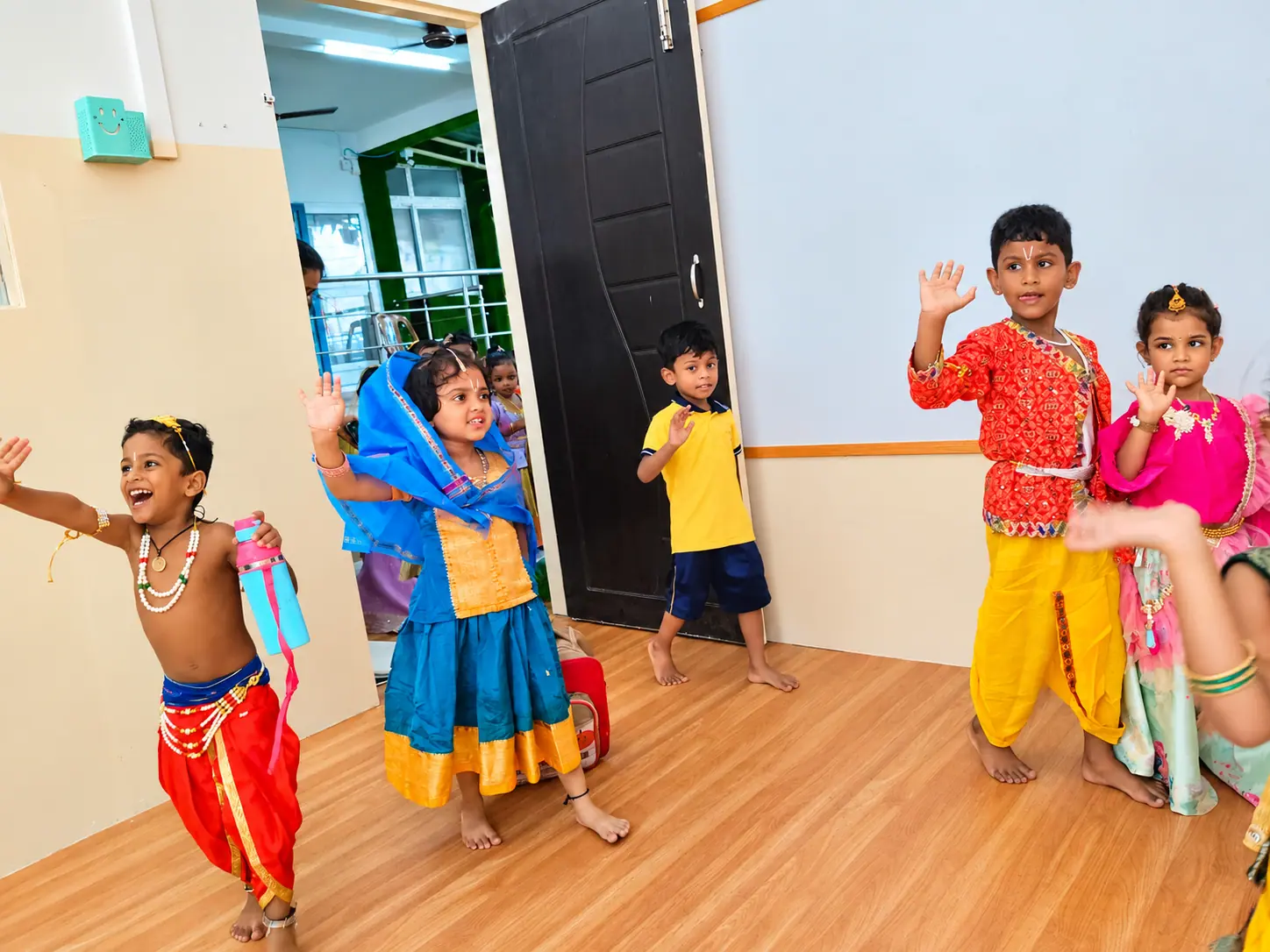 Children in Krishna, Radha and gopi costumes dancing with raised hands on a wooden classroom floor